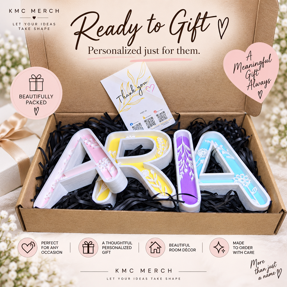 A personalized gift box from KMC MERCH containing large decorative letters spelling 'AURA' in pastel pink, yellow, white, and blue with floral designs. The box is lined with black shredded paper and a small thank-you card.