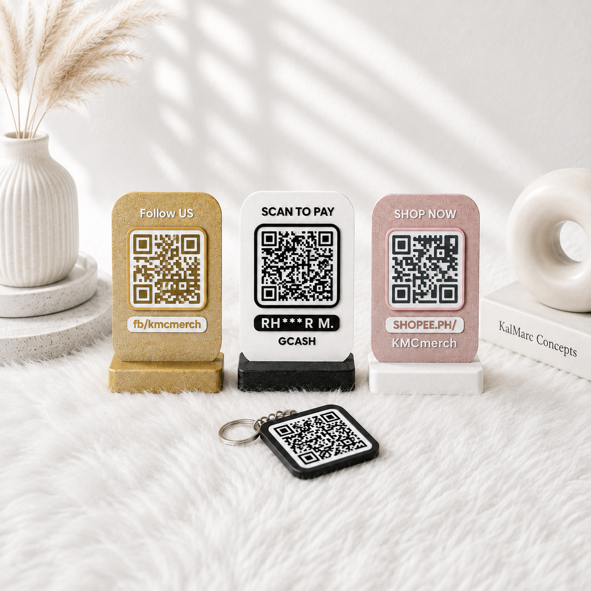 Three QR code payment/display stands in gold, white, and blush pink, plus a matching black keychain with a QR code, arranged on a white faux-fur surface with decorative props in a bright styled setting.